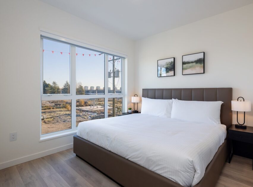 A modern bedroom with a large bed, white bedding, two bedside tables with lamps, two abstract pictures on the wall, and a window with a view of trees and buildings outside.