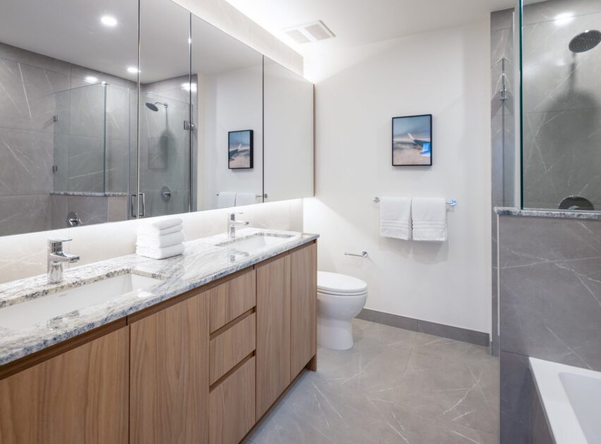Modern bathroom with a double sink vanity, marble countertop, wooden cabinets, a toilet, glass shower, two wall mirrors, white towels, and minimal wall art. The space is bright with neutral tones and sleek fixtures.