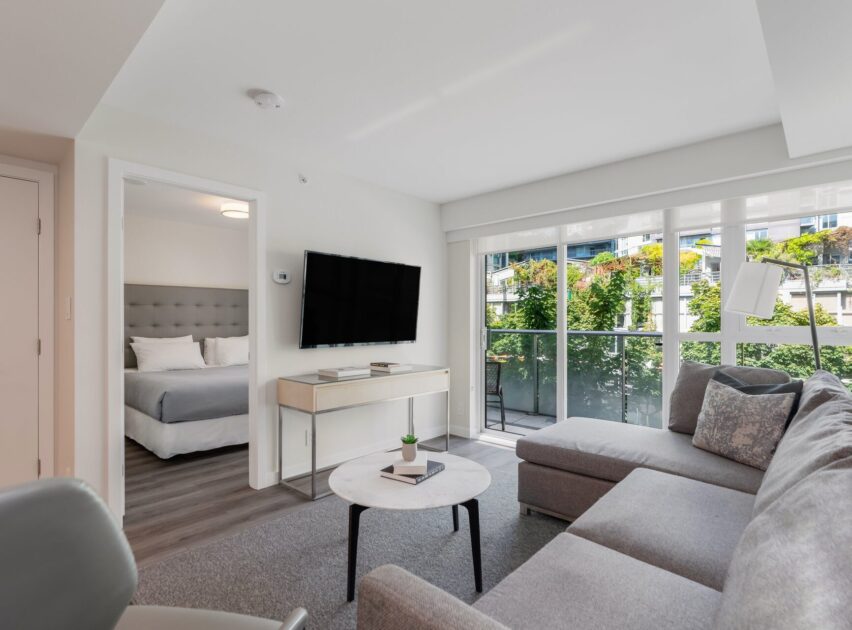 Modern living room with a gray sectional sofa, round coffee table, wall-mounted TV, and large windows. A bedroom with a neatly made bed is visible through an open door. Bright, natural light fills the space.