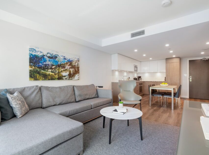 Modern living room with a gray sectional sofa, round white coffee table, and a gray chair. Dining area and kitchen with wood accents in the background. Mountain landscape art on the wall. Bright, open, and minimalist design.