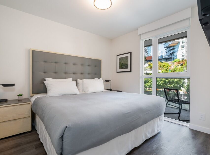 Modern bedroom with a large bed, gray tufted headboard, two nightstands, a window with a view of buildings and greenery, a framed picture on the wall, and a chair outside on a balcony.