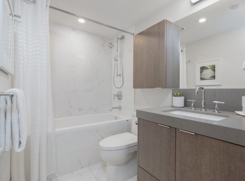 Modern bathroom with a bathtub and shower, gray vanity with sink, wall-mounted cabinets, white marble tile walls and floor, and a small plant on the countertop. Towels hang on a rack next to the tub.