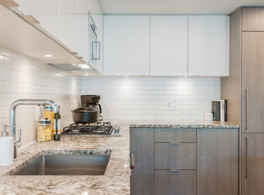 Modern kitchen with granite countertops, stainless steel sink, white subway tile backsplash, white upper cabinets, wooden lower cabinets, a gas stove with cookware, and jars of pasta near the sink.