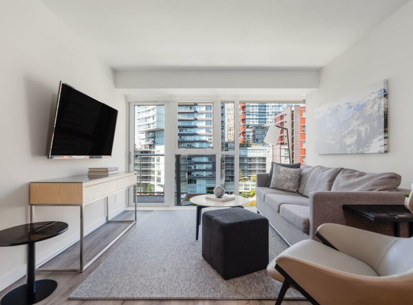 Modern living room with a gray sofa, armchair, ottoman, wall-mounted TV, and large windows offering a city view. Light walls and a neutral rug create a bright, airy atmosphere.