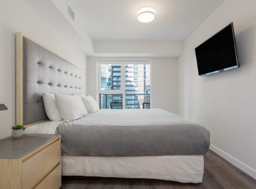 Modern bedroom with a large bed, gray headboard, gray and white bedding, wall-mounted TV, nightstand with a small plant, and a window showing city buildings outside.