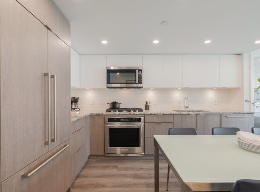 A modern kitchen with light wood cabinets, stainless steel appliances, white upper cabinets, a glass-top dining table with dark chairs, and a bright, minimalist design.