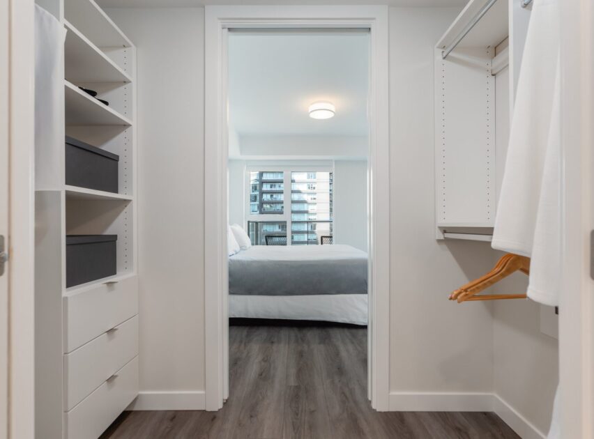 A modern walk-in closet with built-in white shelves and hanging rods leads to a bedroom with a made bed; large windows in the bedroom reveal a view of a neighboring building.