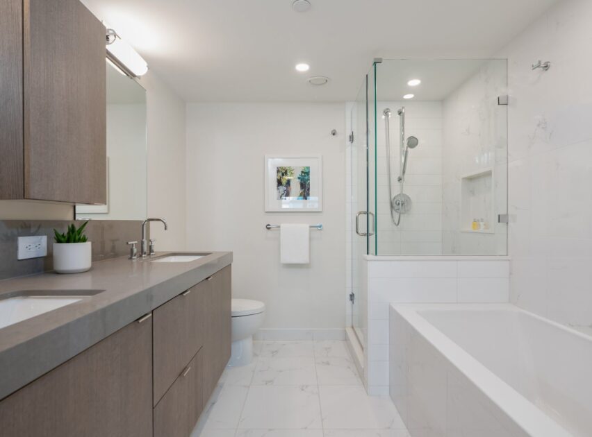 Modern bathroom with a double sink vanity, wall-mounted mirror, potted plant, toilet, glass-enclosed shower, built-in bathtub, and white tile flooring. Soft lighting creates a clean, minimalist look.