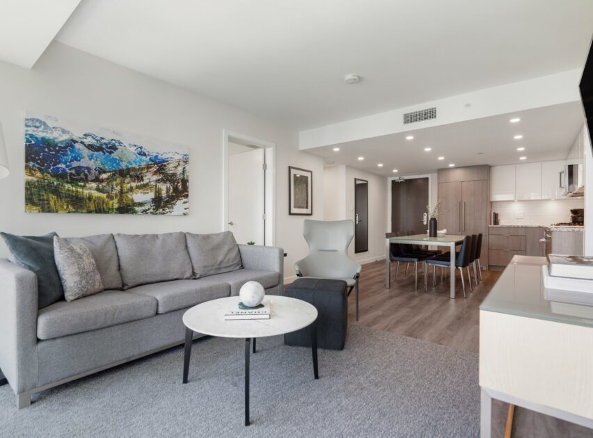 Modern open-concept living room and kitchen with gray sofa, abstract chair, round coffee table, dining area, and wall art depicting mountains. Light wood flooring and neutral tones create a bright, inviting space.