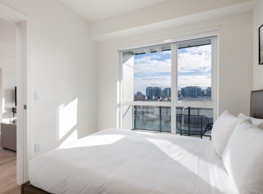 Bright bedroom with a large bed, white bedding, and a big window offering a city skyline and waterfront view; adjacent room and modern decor visible.