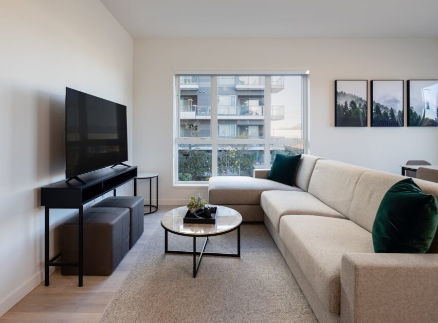 Modern living room with a beige sectional sofa, dark green pillows, round coffee table, TV on a stand, and large window showing a balcony and apartment building outside. Three forest-themed prints hang on the wall.