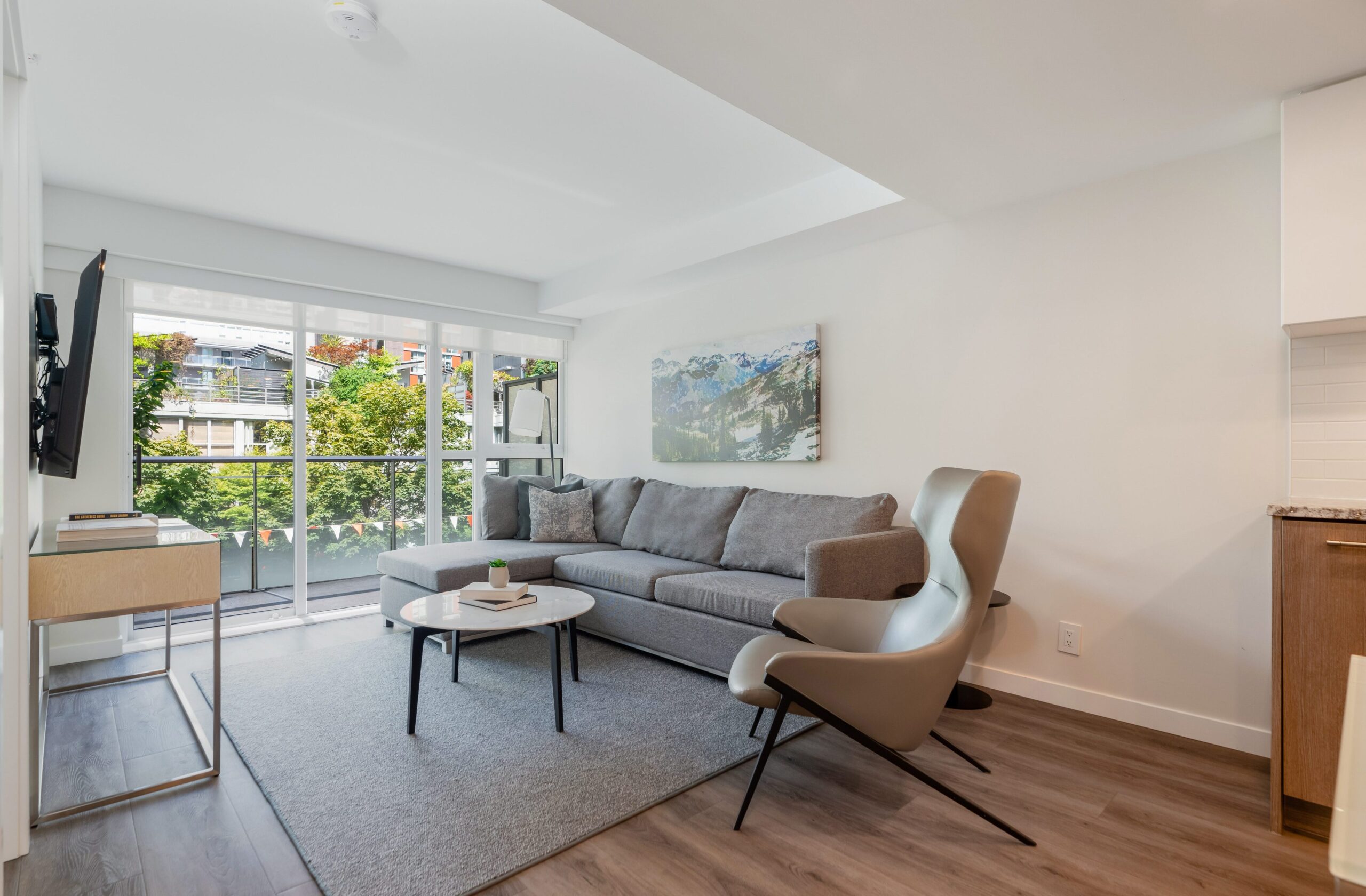 A modern living room with a gray sofa, coffee table, and armchair, featuring large windows that overlook trees and buildings. The space is bright with natural light and decorated in neutral tones.