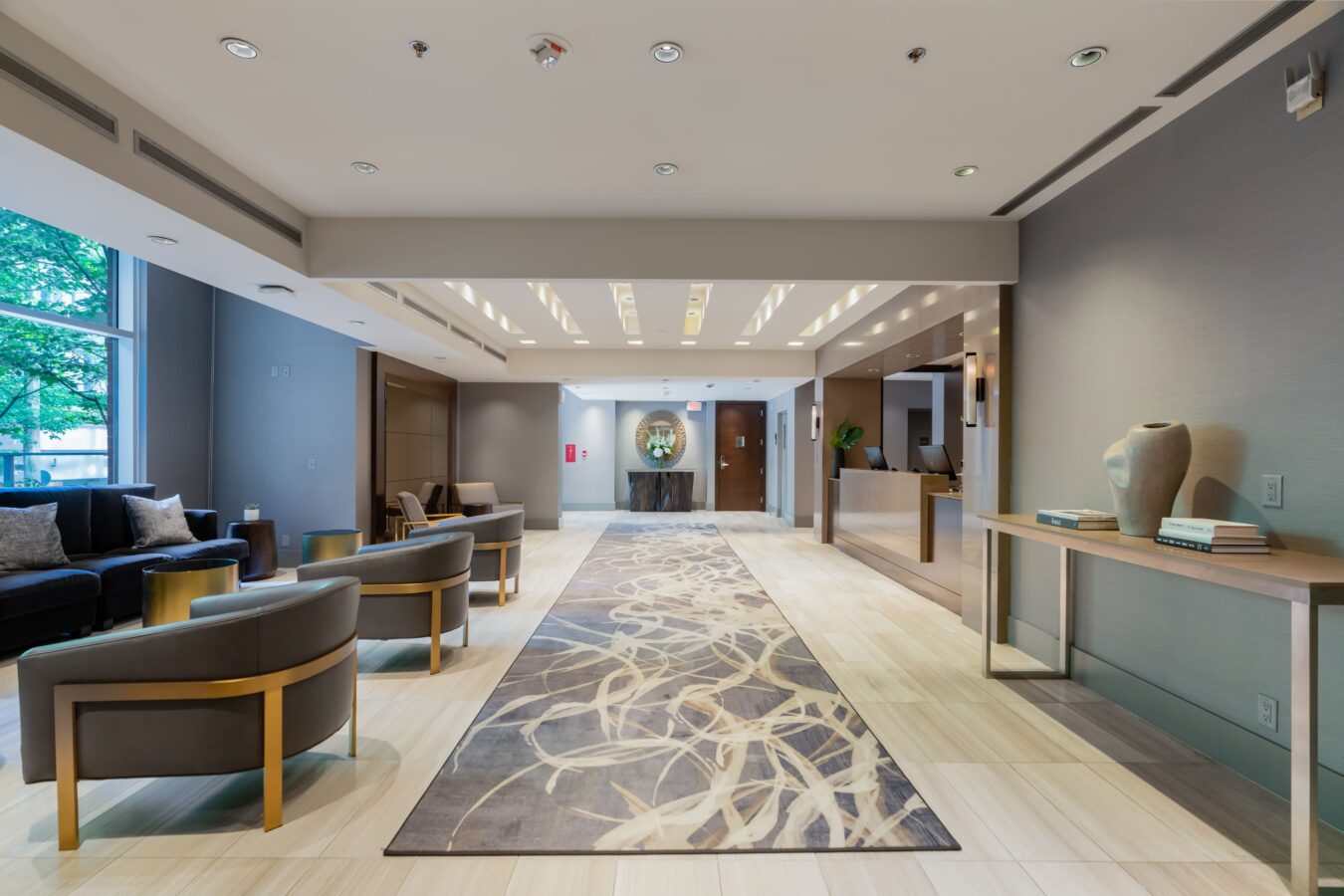 Modern, spacious hotel lobby with neutral tones, two seating areas, a long decorative rug, large windows on the left, a reception desk on the right, and a console table with vases against the wall.