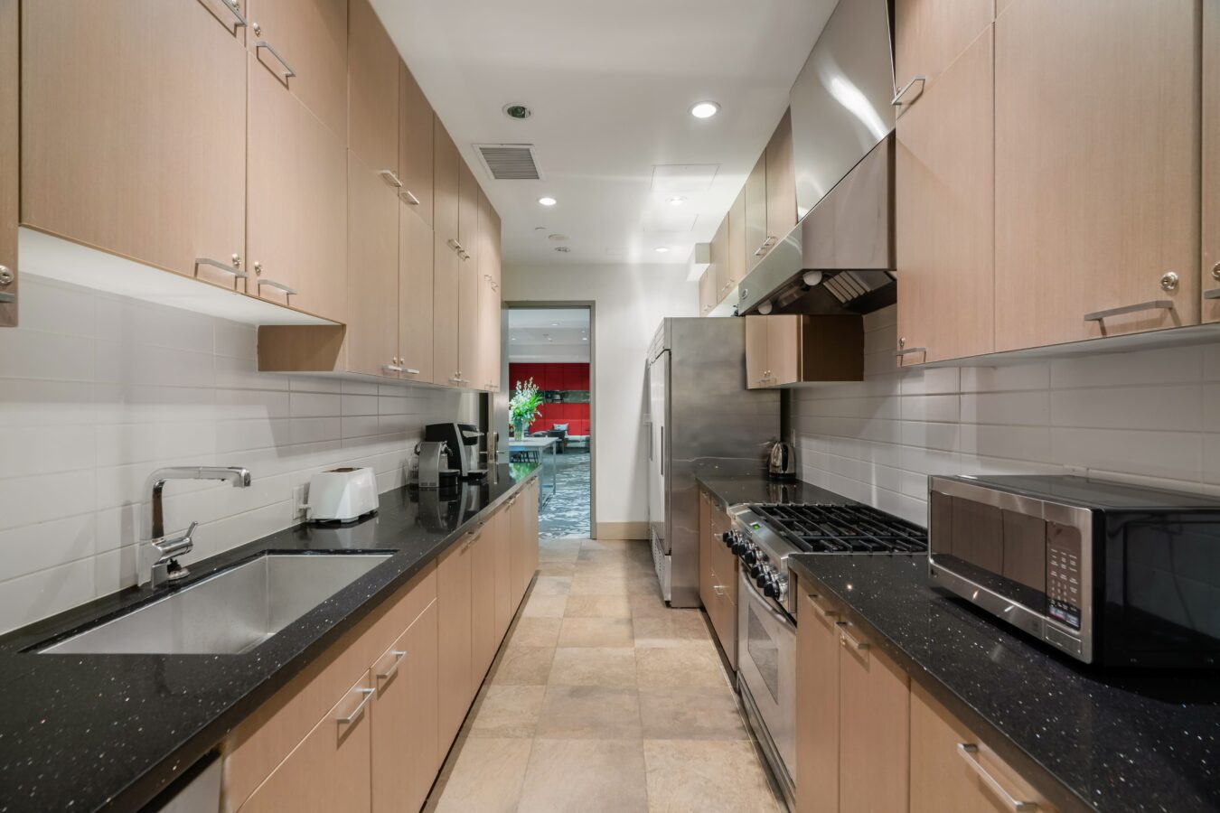 Modern galley kitchen with light wood cabinets, black countertops, stainless steel appliances, microwave, sink, gas stove, and tile floor. A bright dining area is visible at the end of the room.