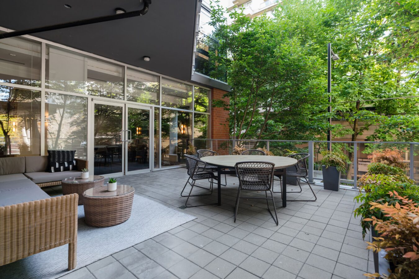A modern patio with wicker furniture, a round dining table with chairs, potted plants, and trees, next to a building with large glass doors and windows.