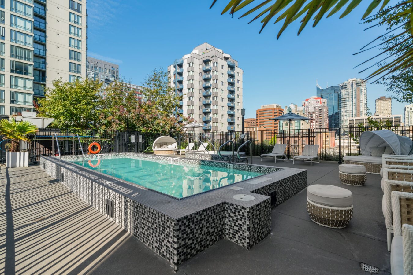 A rooftop pool surrounded by lounge chairs and round tables, with city skyscrapers and trees in the background under a clear blue sky.