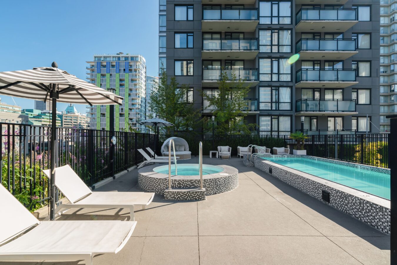 Rooftop pool area with lounge chairs, a round hot tub, and a slim rectangular swimming pool next to a modern apartment building, surrounded by a black fence and city buildings in the background.