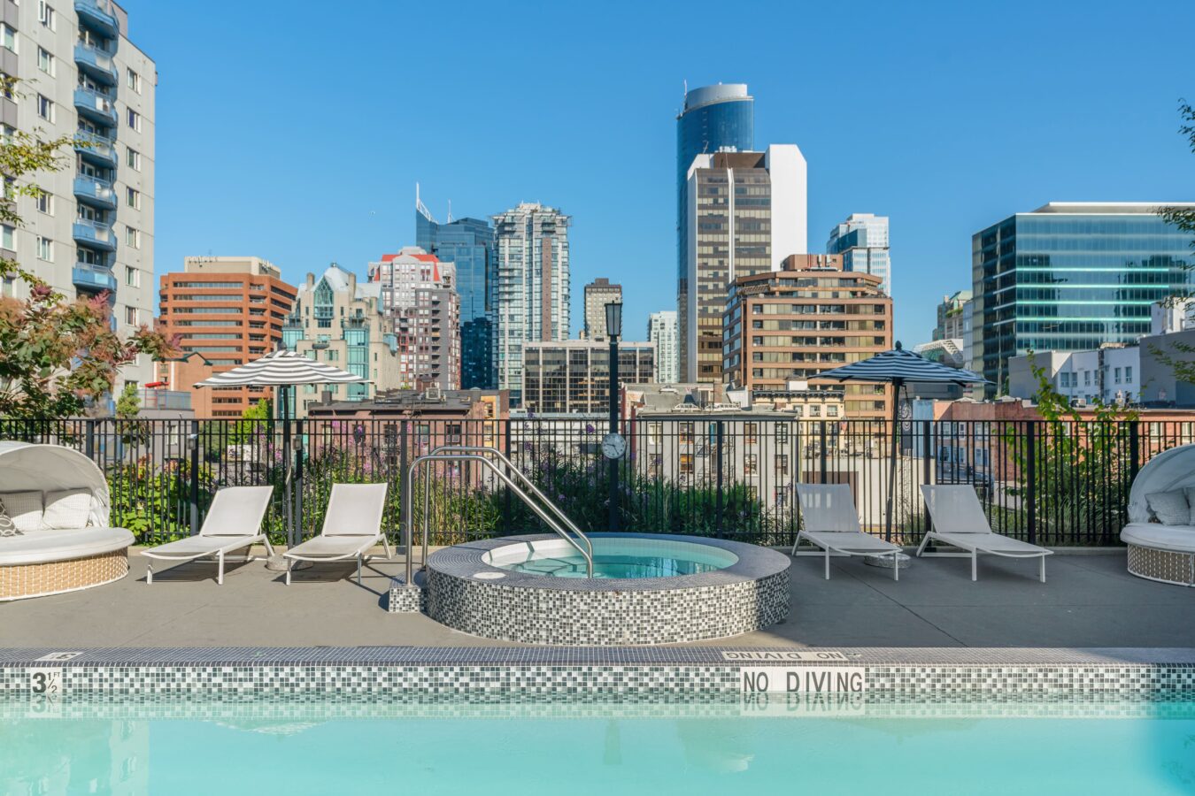 A rooftop pool and hot tub surrounded by lounge chairs and umbrellas, with a city skyline of modern high-rise buildings in the background under a clear blue sky.