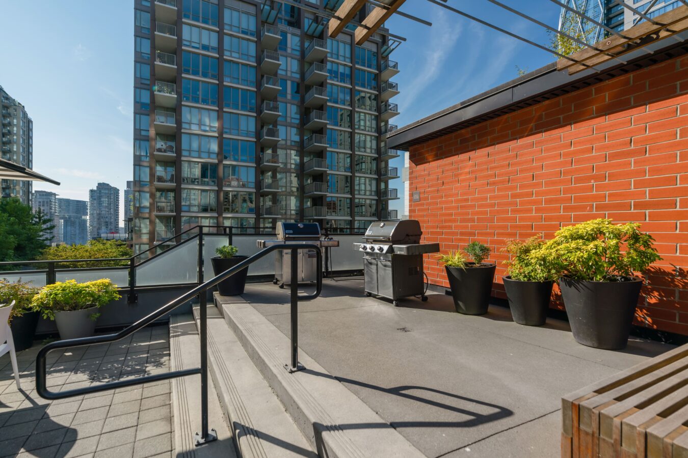 Outdoor terrace with potted plants, a barbecue grill, and a brick wall, overlooking modern high-rise buildings on a sunny day. Steps and black railings lead up to the grilling area.