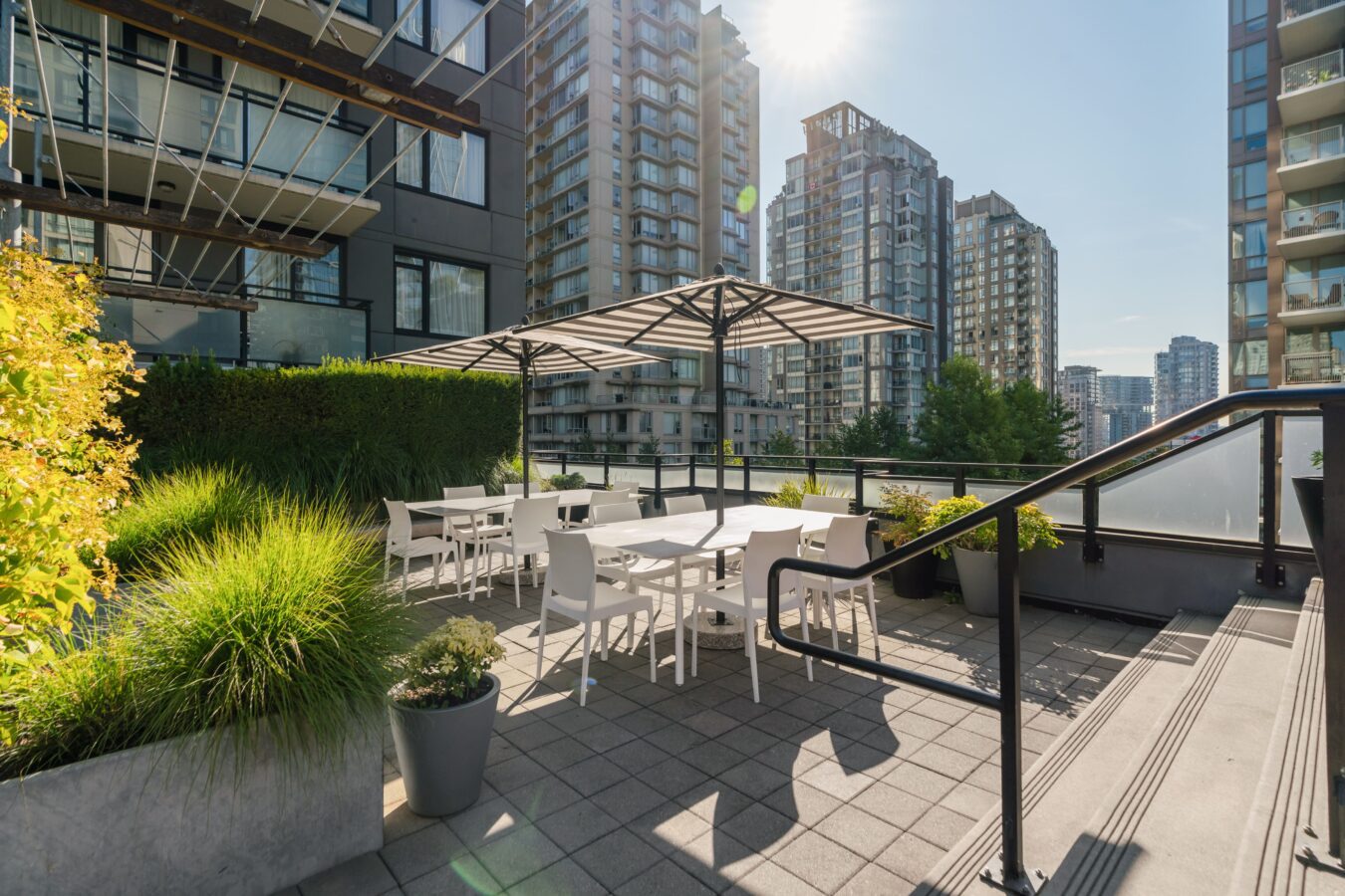 Sunny rooftop patio with white tables and chairs under large umbrellas, surrounded by potted plants and greenery, with tall modern buildings in the background.