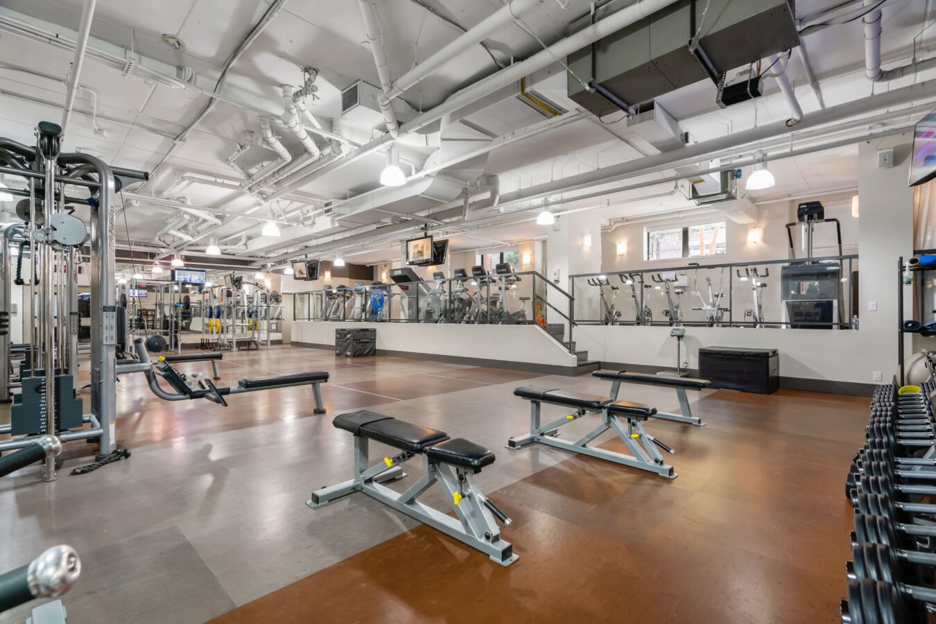 Spacious, modern gym with weight benches, strength machines, free weights, and cardio equipment on an upper level. The gym has bright lighting, polished floors, and a clean, open layout.