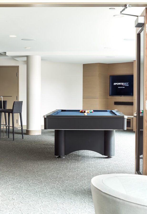 A modern game room with a pool table set up for a game, a flat-screen TV mounted on the wall displaying SPORTSNET, and bar stools at a tall table in the background. The space is bright with natural light.