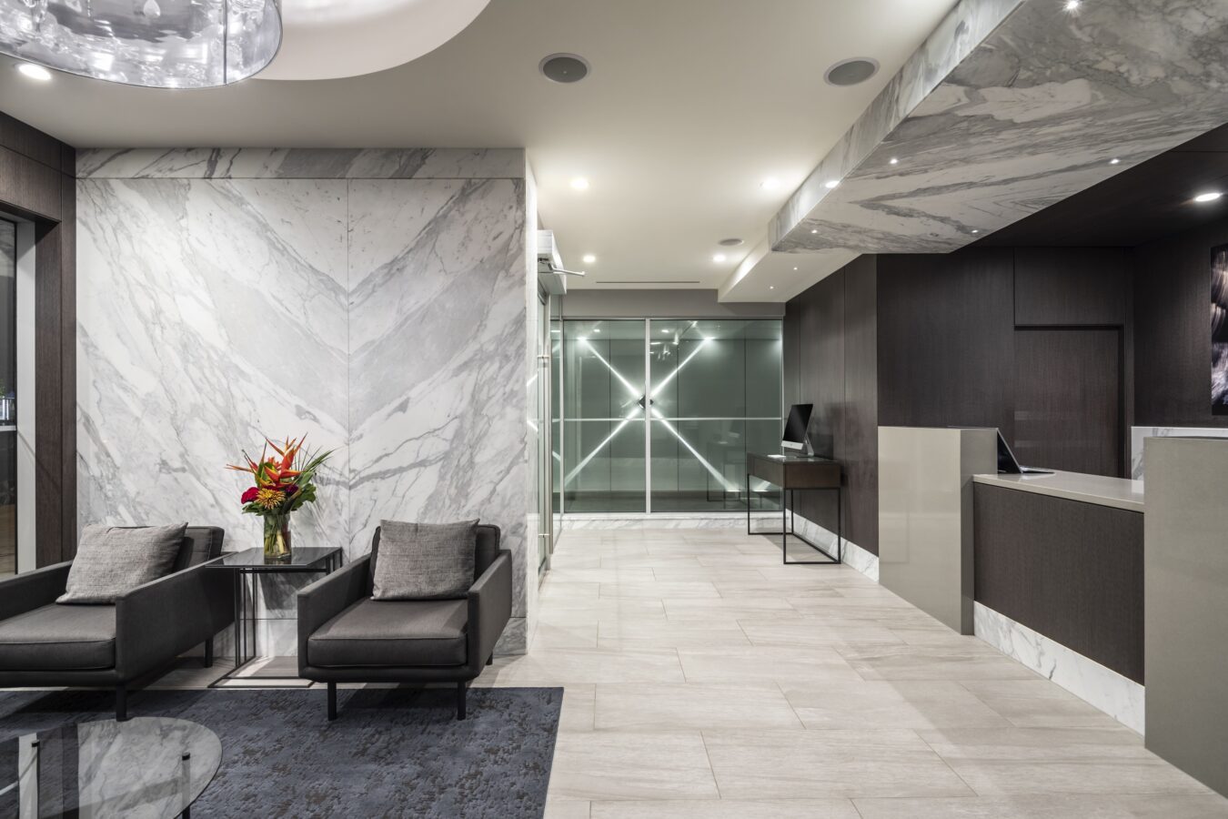 Modern office lobby with marble walls and floors, two black armchairs with gray cushions, a glass coffee table with a flower arrangement, and a sleek reception desk under bright lighting.