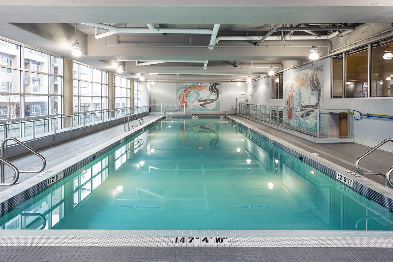Indoor swimming pool with turquoise water, surrounded by metal railings and large windows. A mural of fish decorates the far wall. Overhead lights illuminate the space, and safety markings are visible on the pool edge.