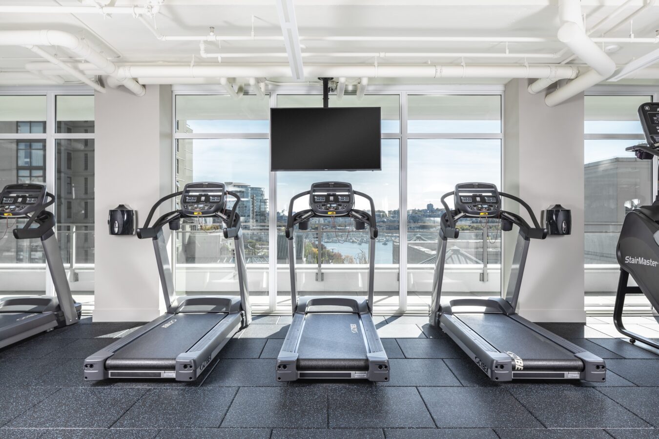 A modern gym with three treadmills facing large windows, a flat-screen TV mounted above them, and an exercise bike on the right; city buildings and water are visible outside.