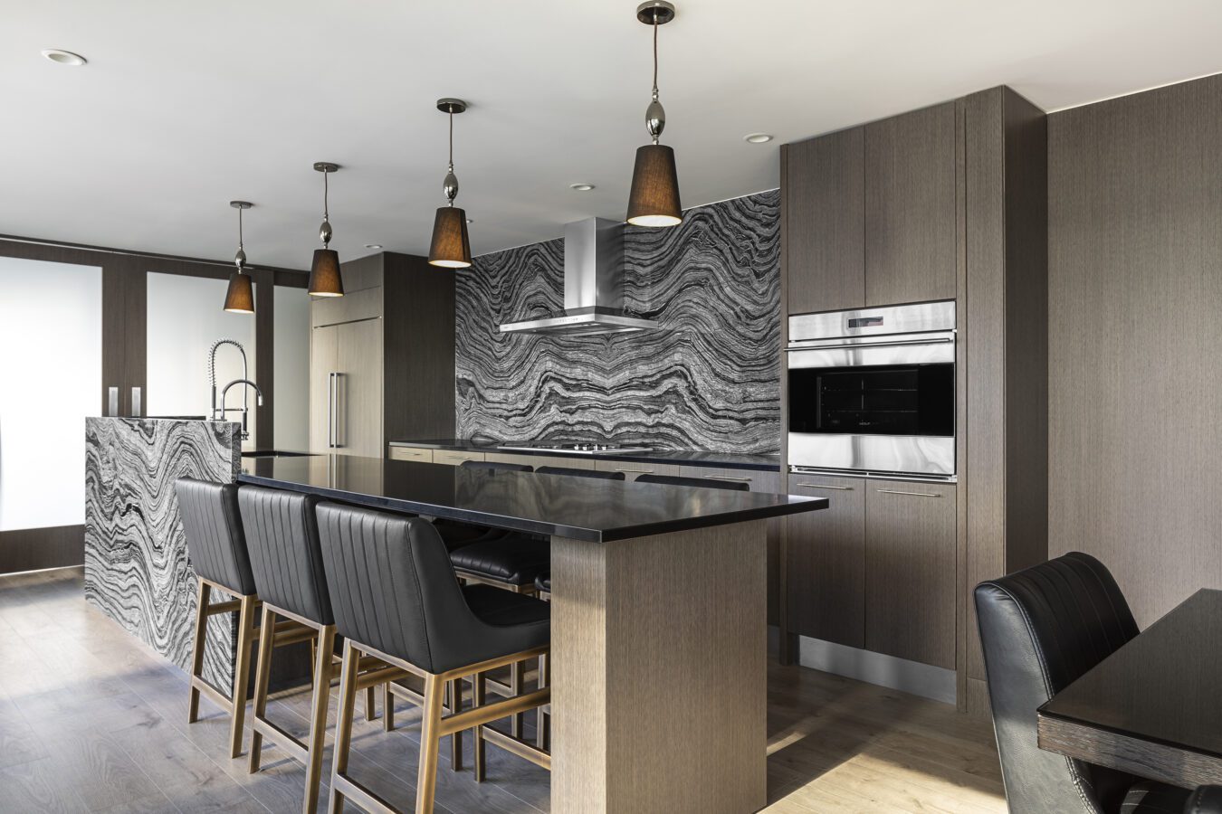 Modern kitchen with dark wood cabinets, black countertops, and a striking black-and-white marble backsplash and island. Four pendant lights hang above the island, and there are black leather chairs and built-in stainless steel appliances.