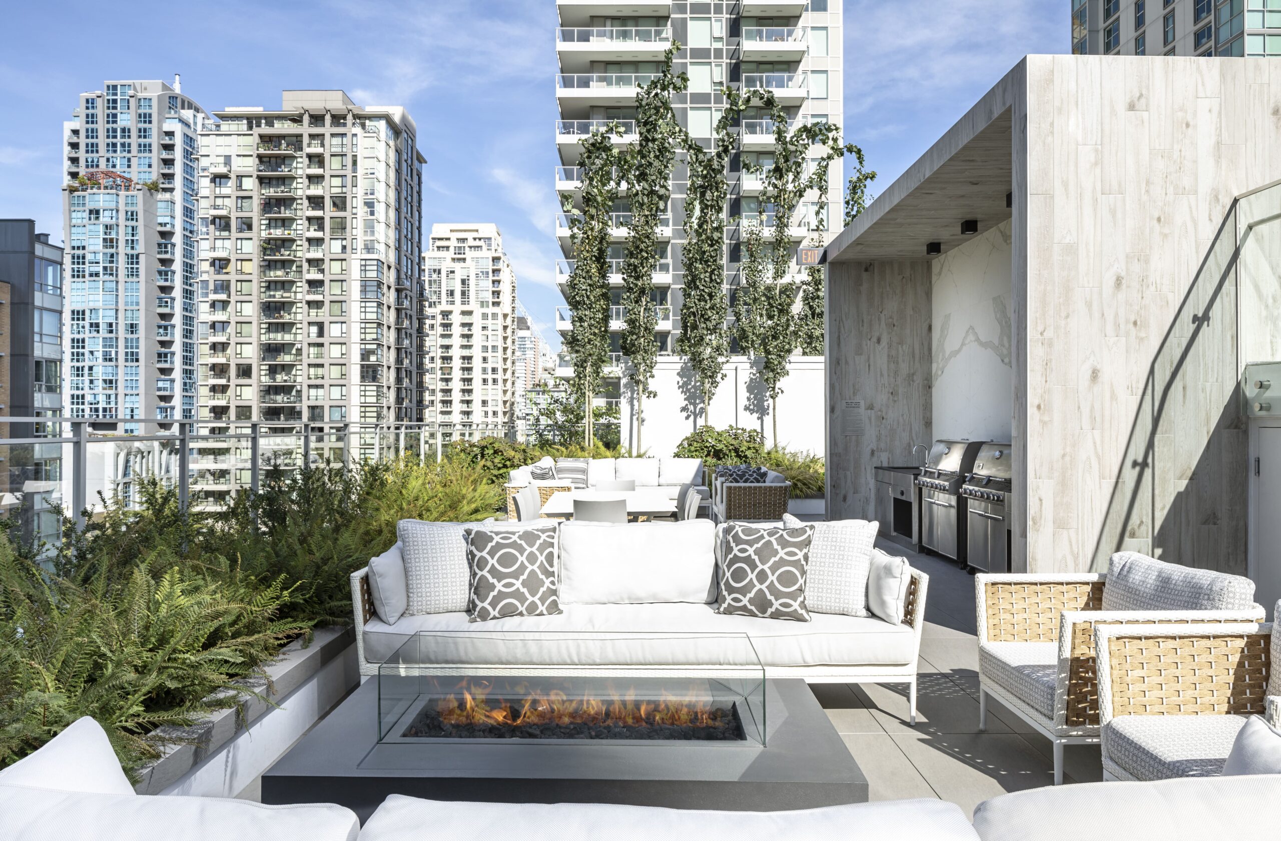 Modern rooftop patio with white seating, a glass fire pit, outdoor dining area, built-in grill, greenery, and tall city buildings in the background under a blue sky.