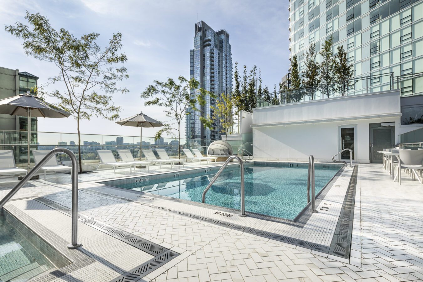 A modern rooftop swimming pool area with lounge chairs, umbrellas, and glass railings, surrounded by tall buildings and greenery on a sunny day.