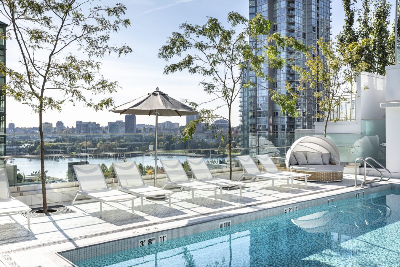 A modern rooftop pool with lounge chairs, an umbrella, and a round daybed, surrounded by glass railings and trees. High-rise buildings and a city skyline with a river are visible in the background.