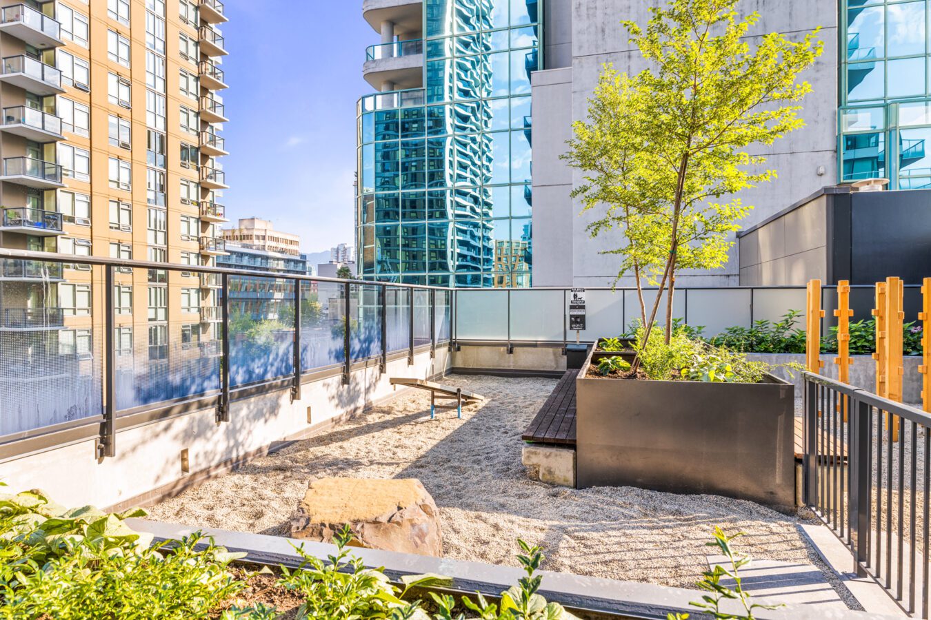 A modern rooftop terrace with a sand play area, a wooden bench, a large planter with a small tree, and surrounding tall glass buildings under a clear blue sky.