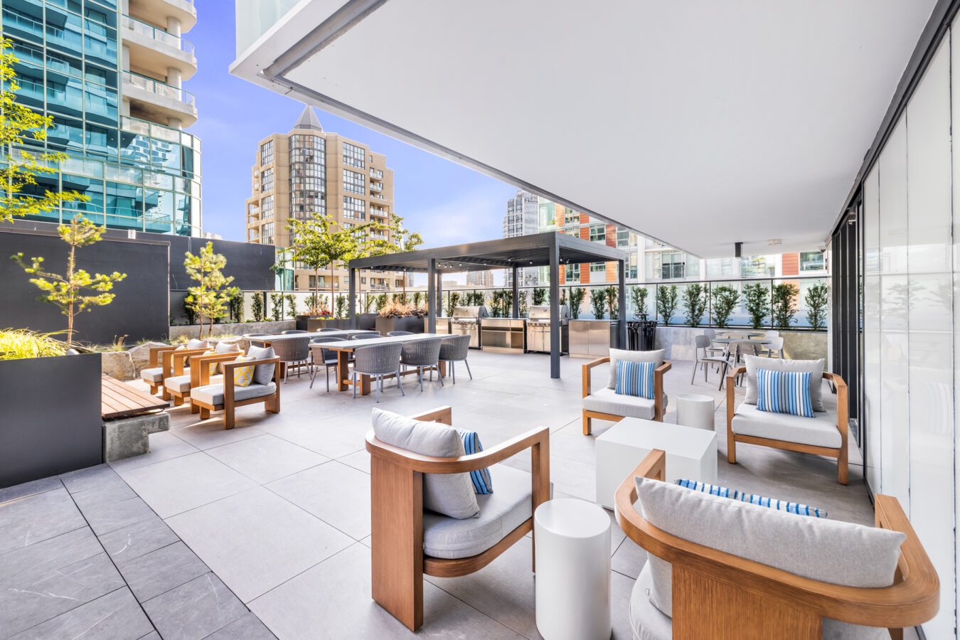 Modern outdoor lounge area on a rooftop with wooden chairs, cushioned seating, tables, and potted plants, surrounded by glass railings and city buildings under a clear blue sky.