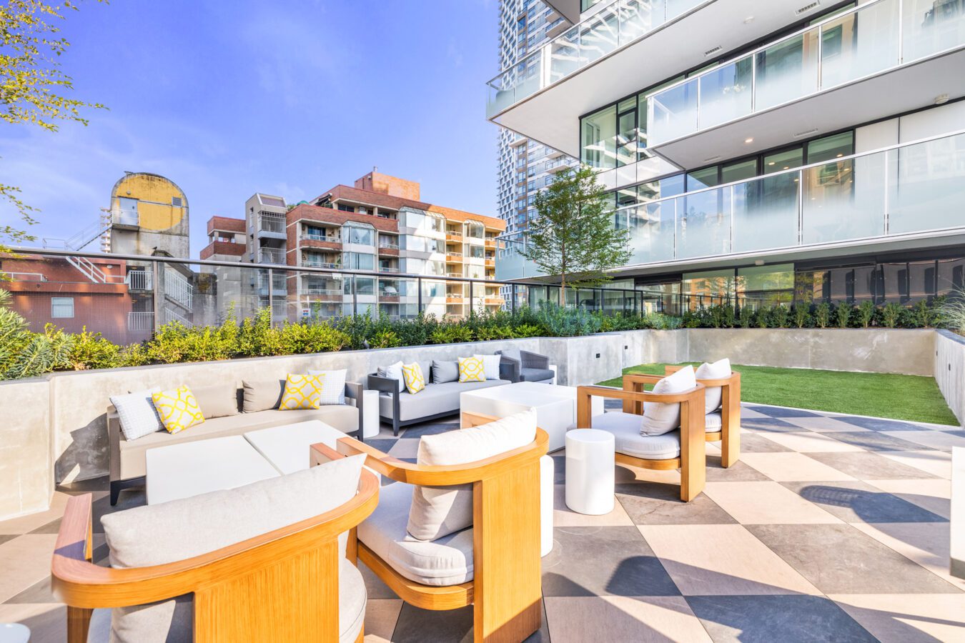 Modern outdoor lounge area with cushioned chairs and sofas on a checkered patio, bordered by greenery, next to a glass-walled building with city apartment buildings in the background under a clear blue sky.