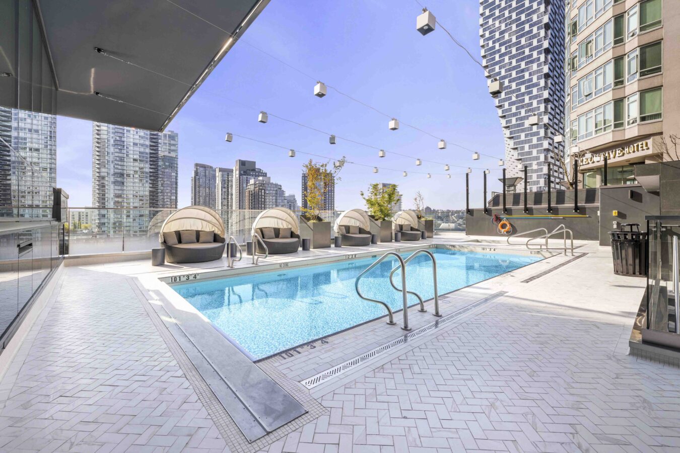 Rooftop swimming pool surrounded by lounge chairs and modern buildings, with string lights overhead and a clear blue sky in the background.