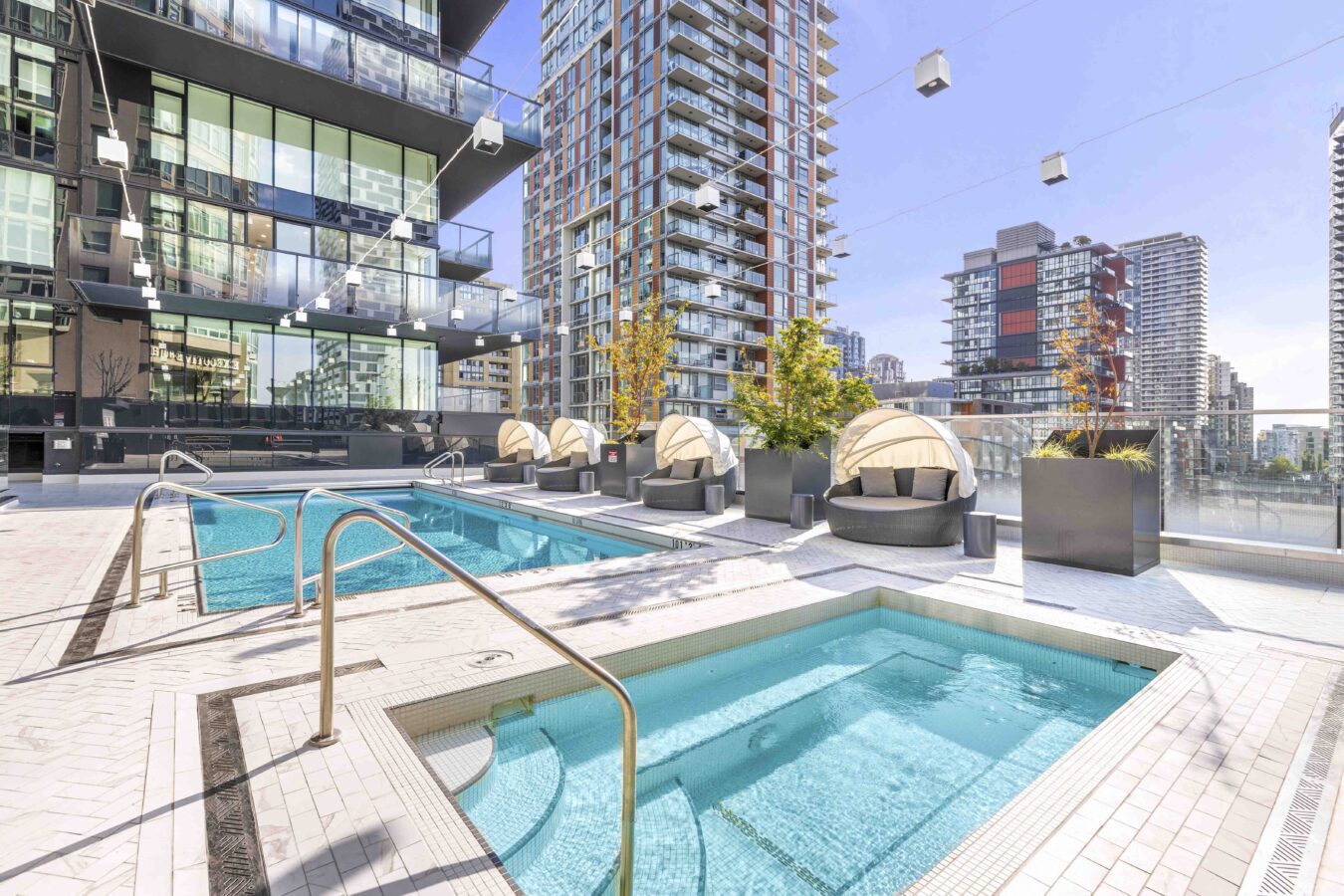 Modern rooftop pool area with a hot tub, lounge chairs, cabanas, and tall buildings in the background under a clear blue sky.
