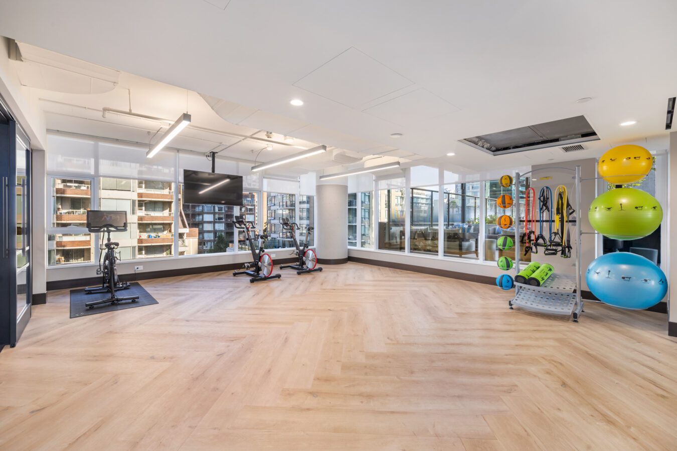 A modern, bright gym with large windows, two stationary bikes, a TV, colorful exercise balls on the wall, and various fitness equipment neatly organized in a spacious room with light wooden floors.