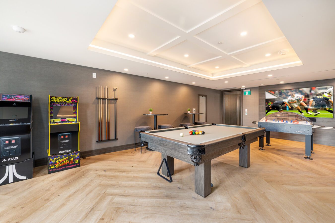 A modern game room with a pool table, arcade game machines, a foosball table, cues on a wall rack, and a large TV showing a football game. The room has wood floors and neutral-colored walls.