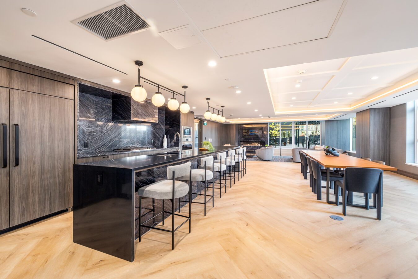 Modern communal kitchen and dining area with wood flooring, a long black marble island with barstools, pendant lights, and large windows providing natural light. Lounge area and dining table visible in the background.