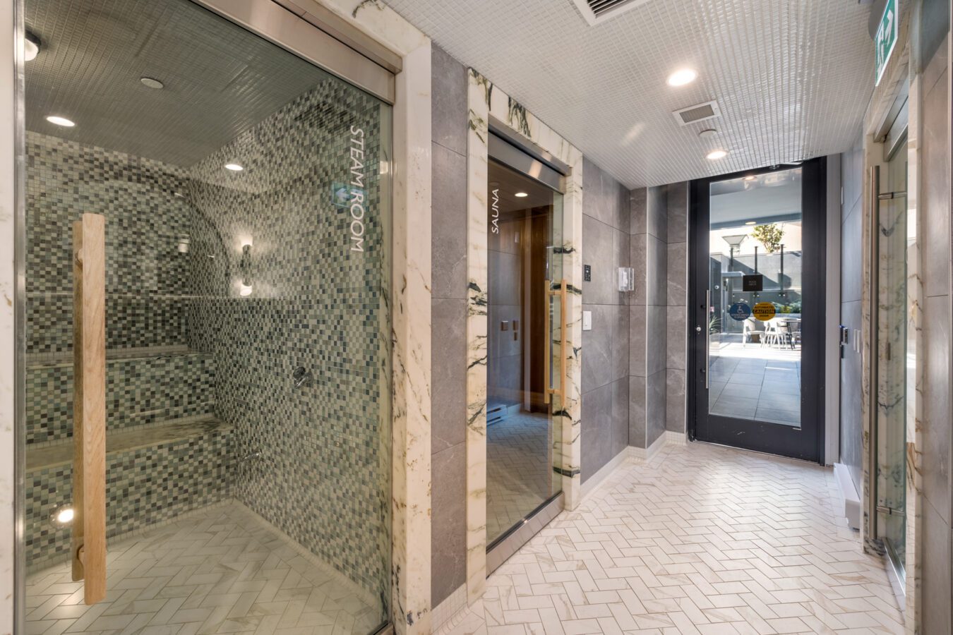 A modern spa hallway with a glass steam room on the left, a sauna with wooden benches in the center, and a door leading outside to a pool area in the background. The floor is tiled in a herringbone pattern.