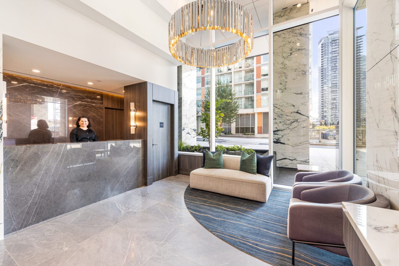 Modern lobby with marble floors and walls, large windows, a chandelier, a curved sofa with pillows, two armchairs, and a receptionist standing behind a sleek dark reception desk. Urban buildings visible outside.