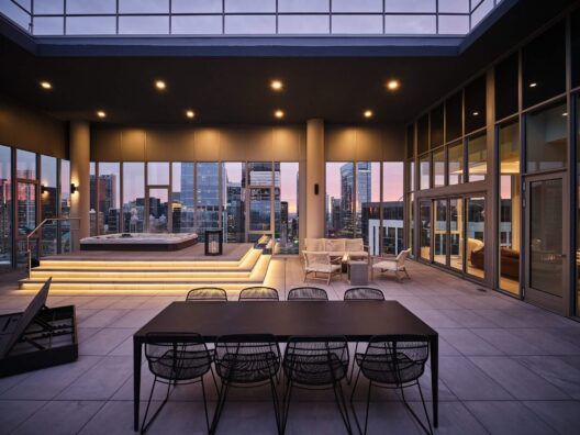 Modern rooftop terrace at sunset with a dining table, chairs, lounge seating, and a lit-up hot tub. Large glass windows showcase a city skyline with tall buildings in the background.