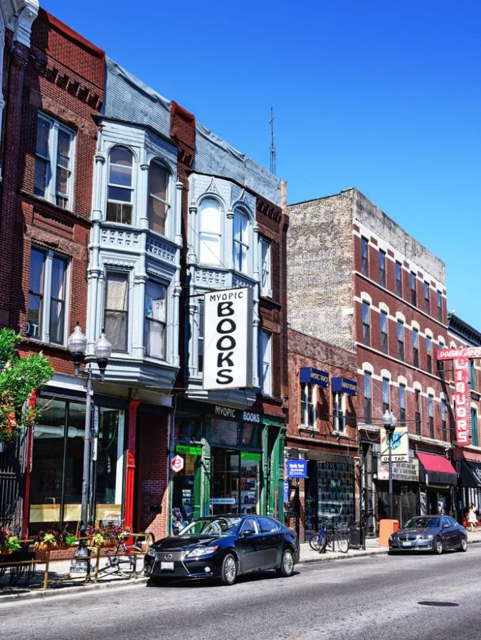 A city street lined with historic brick buildings features a prominent bookstore called Myopic Books. Cars and bicycles are parked along the sidewalk under a clear blue sky.