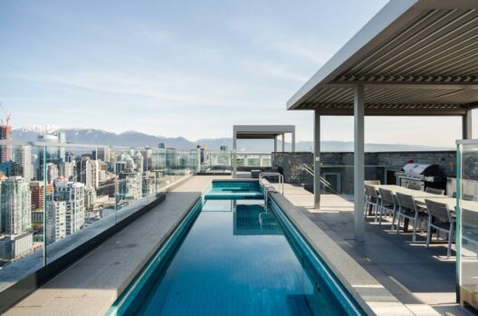 A modern rooftop features a narrow swimming pool, lounge chairs, and a shaded dining area with a grill, overlooking a city skyline and distant snow-capped mountains under a clear sky.