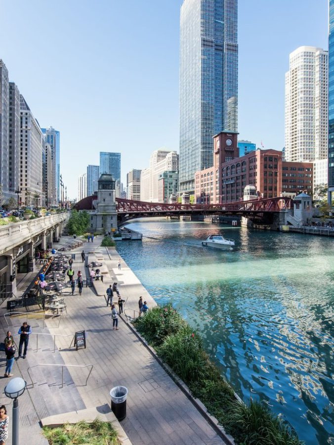 A riverside walkway in downtown Chicago with people walking, tall modern buildings, a red bridge over the Chicago River, and a boat traveling on the water under a clear blue sky.