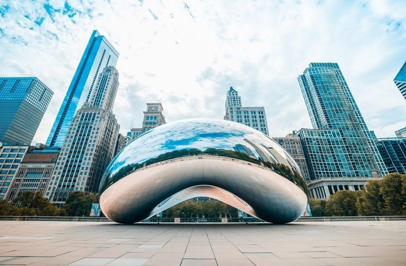 Cloud Gate, also known as The Bean, is a large, reflective sculpture in Chicago’s Millennium Park, surrounded by tall city buildings under a partly cloudy sky.
