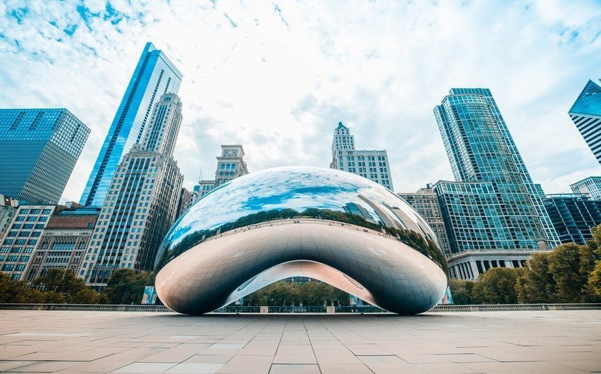 The photo shows Cloud Gate, a large reflective sculpture in Chicago’s Millennium Park, with city skyscrapers and a partly cloudy sky in the background.
