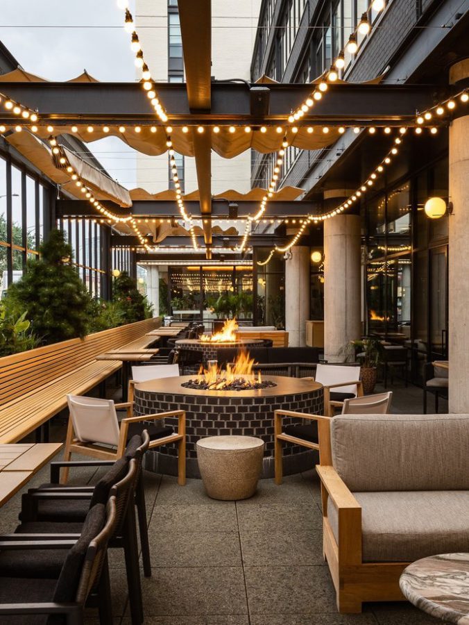 A modern outdoor patio with string lights overhead, a round brick fire pit in the center, wooden benches, cushioned chairs, and green plants along the sides, creating a cozy and inviting atmosphere.